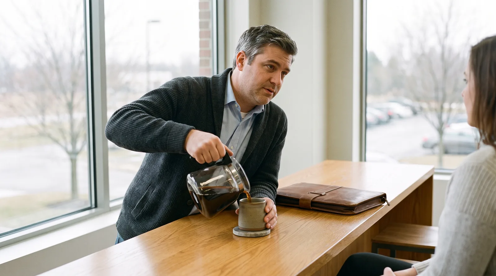 A man in a cardigan pours coffee from a glass carafe into a ceramic mug on an oak counter in an office kitchen.
